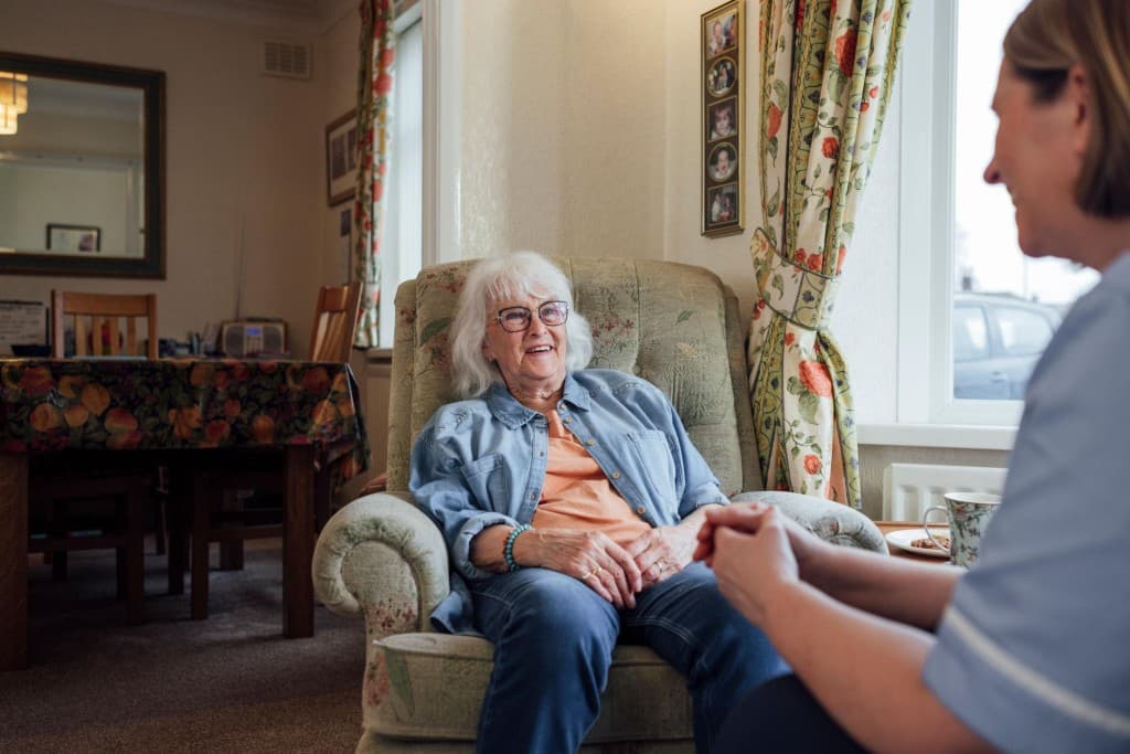 An elderly woman smiling warmly while receiving friendly companionship from a professional caregiver in a comfortable home setting